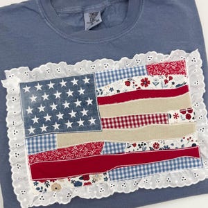 Puede incluir: Una sudadera azul con un diseño de bandera americana de patchwork. La bandera incluye estrellas blancas sobre un campo de mezclilla azul, rayas rojas y blancas, y un borde de encaje blanco. La etiqueta dice "Comfort Colors".