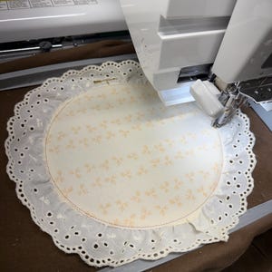 May include: A cream-colored, circular doily with a floral pattern is being embroidered by a white sewing machine. The doily has a scalloped, eyelet lace edge. The machine's needle is positioned over the fabric, ready to stitch.