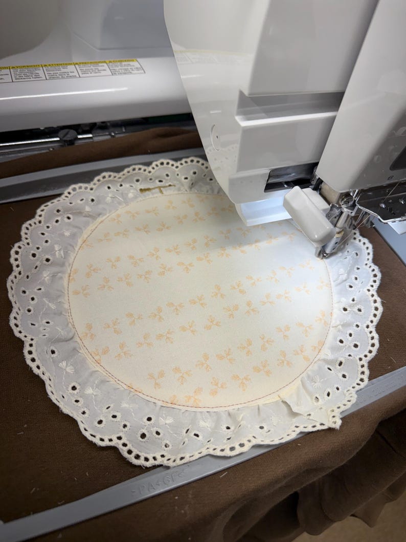 May include: A cream-colored, circular doily with a floral pattern is being embroidered by a white sewing machine. The doily has a scalloped, eyelet lace edge. The machine's needle is positioned over the fabric, ready to stitch.
