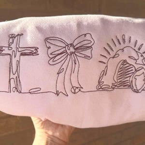 May include: A light pink fabric featuring dark brown embroidered line art of a cross, a bow, and an empty tomb with a shield. The fabric is held by a hand.