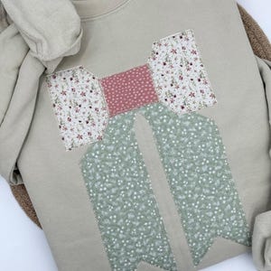 May include: A beige sweatshirt with a large bow design. The bow features a pink center with white polka dots, floral print on the top sections, and green floral print on the bow tails. The sweatshirt is folded.