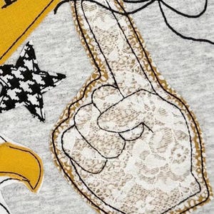 May include: A gray fabric with a large foam finger applique. The foam finger is white lace with a gold border and black outline. Other appliqued elements include a black and white star, a yellow triangle, and a white eagle head.