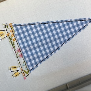 Könnte beinhalten: Ein dreieckiger Wimpel mit blau-weißem Vichy-Muster. Der Wimpel ist mit schwarzen Nähten und einem floralen Rand versehen. Zwei gelb-weiße Laschen ragen aus dem Wimpel heraus.