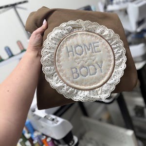May include: A brown sweatshirt with a circular embroidered design. The design features the words "HOME BODY" in blue stitching, surrounded by a white lace trim. The sweatshirt is held by a person.