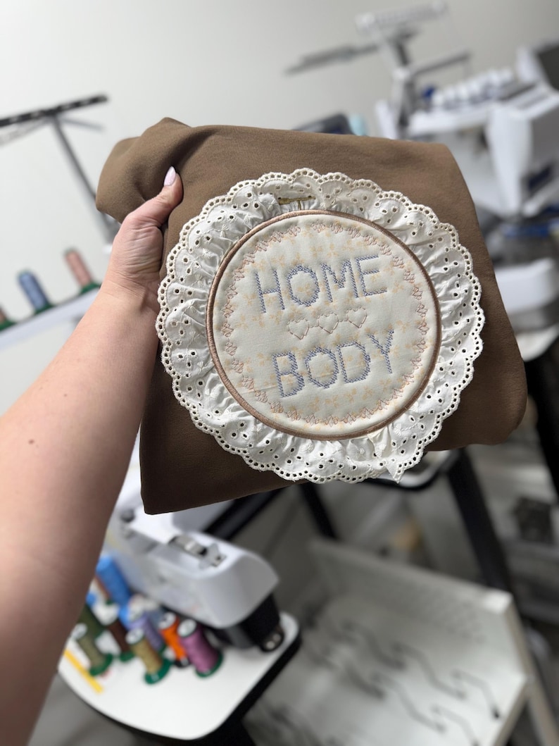 May include: A brown sweatshirt with a circular embroidered design. The design features the words "HOME BODY" in blue stitching, surrounded by a white lace trim. The sweatshirt is held by a person.