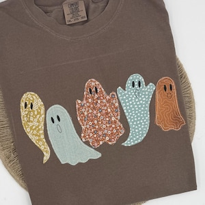 Puede incluir: Una camiseta marrón de manga corta con cinco aplicaciones de tela en forma de fantasma con varios patrones, incluyendo flores, lunares y texturas sólidas. La etiqueta Comfort Colors en el cuello muestra claramente la talla 3XL-3TG-3EG.