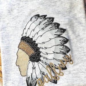 May include: A grey t-shirt with an embroidered Native American headdress design. The headdress features black, white, and gold detailing, and the word "gallery" is embroidered in gold script.
