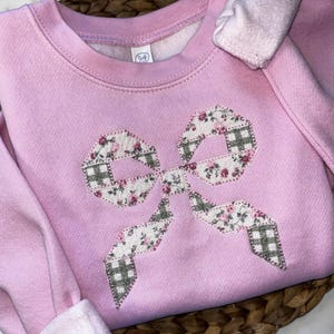 May include: A light pink sweatshirt with a large bow applique. The bow is made of floral and plaid patterned fabric. The sweatshirt has long sleeves and a crew neck.