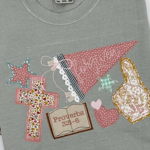 May include: A gray Comfort Colors t-shirt with various appliqued patches. Patches include a cross, a pennant with "forgiven" on it, stars, a heart, a foam finger, and a book with "Proverbs 3:5-6" on it.