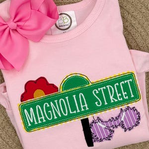 May include: A light pink toddler's shirt with a Magnolia Street sign applique. The sign is green with white lettering and a yellow border. A large pink bow is attached to the top left. A red flower and purple accents complete the design.