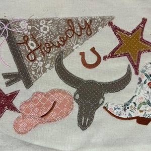 May include: A fabric banner with the word "Howdy" embroidered in brown thread, alongside a horseshoe, star, cowboy hat, bull skull, and a floral-patterned cowboy boot. The design features a color palette of brown, pink, and cream.