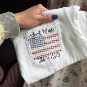 May include: White t-shirt with a pocket featuring a cross-stitched American flag design. The flag is red, white, and blue, with the words "God bless the USA" embroidered above and below the flag. The shirt is held by a person.