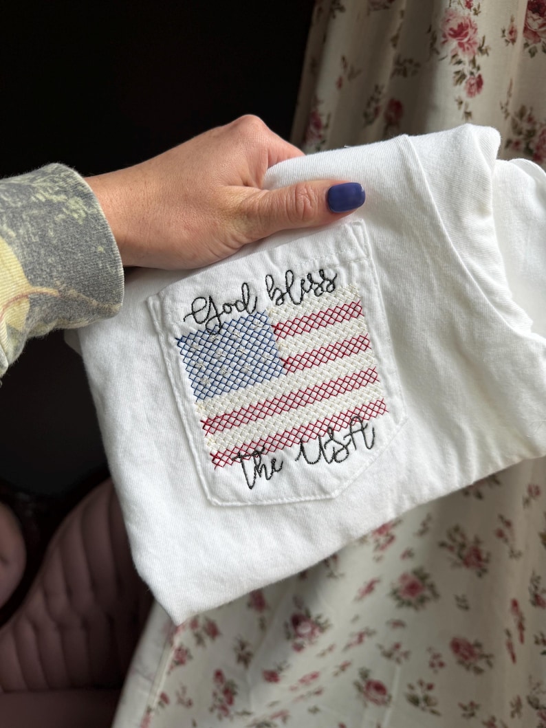 May include: White t-shirt with a pocket featuring a cross-stitched American flag design. The flag is red, white, and blue, with the words "God bless the USA" embroidered above and below the flag. The shirt is held by a person.