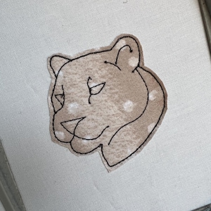 May include: Embroidered patch featuring a lioness head in profile. The lioness is tan with white polka dots and outlined in black thread. The patch is set in an embroidery hoop.