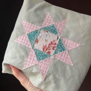 May include: A pale green cushion featuring a star-shaped appliqué. The star is made from pink and teal fabric, with a floral pattern at the centre. The cushion appears to be made from linen or a similar soft material.