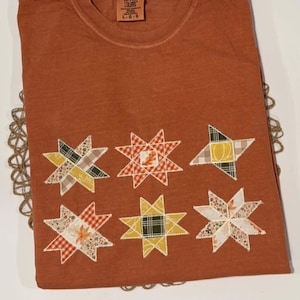 May include: A rust-colored t-shirt featuring six appliqued star shapes arranged in two rows. Each star is constructed from various fabric patches in plaid, gingham, and floral patterns in shades of orange, yellow, green, and white.