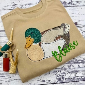 May include: A tan coloured sweatshirt featuring an embroidered duck design. The duck has a green head, yellow beak, and various tan and brown patches. The name "Blaise" is embroidered in green script.