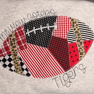 Könnte beinhalten: Ein hellgraues Sweatshirt mit einer aufgestickten Football-Applikation aus verschiedenen Stoffmustern wie rot-schwarzem Schottenkaro, Punkten, Streifen und Leopardenmuster. Der Text Mt. View Gotebo ist darüber und Tigers darunter eingestickt.