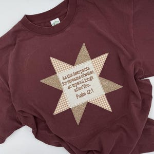 Könnte beinhalten: Ein kastanienbraunes T-Shirt mit kurzen Ärmeln und einer zentralen sternförmigen Applikation. Der Stern wechselt zwischen einem beigen Vichy-Muster und einem gesprenkelten beigen Stoff. Das mittlere Quadrat zeigt den Text: As the deer pants for streams of water, so my soul longs after You. Psalm 42:1.