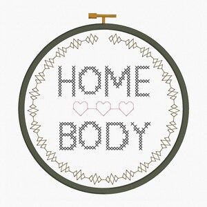 May include: A cross-stitch embroidery hoop with the words "HOME BODY" in black thread, with three pink hearts. The design is framed by a green hoop and a decorative border. A wooden clasp is at the top.