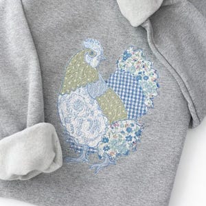 Peut inclure: Un sweat-shirt gris avec un appliqué de coq patchwork coloré. Le coq présente des motifs floraux et vichy bleus, verts et blancs. Les manches ont un poignet doublé de polaire gris clair. Le motif est cousu sur le devant du sweat-shirt.