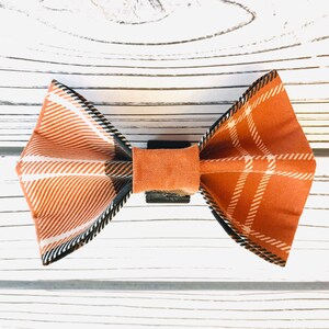 May include: A brown and white plaid bow tie with black trim. The bow tie is tied with a brown fabric band.