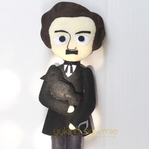 May include: A felt doll of Edgar Allan Poe holding a black felt raven. The doll is wearing a black suit with a white shirt and a black tie. The doll has brown hair and a mustache.