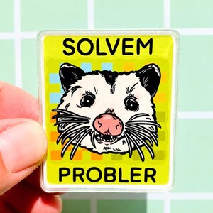 May include: A rectangular acrylic pin featuring a black and white illustration of an opossum face with a pink nose. The background is a yellow grid pattern with the text SOLVEM at the top and PROBLER at the bottom in bold black capital letters.