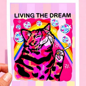 May include: A rectangular art print featuring a pink and black tabby cat with eyes closed, holding a cigarette. The cat is surrounded by colorful bubbles and a rainbow against a checkered background. The text LIVING THE DREAM is printed at the top.