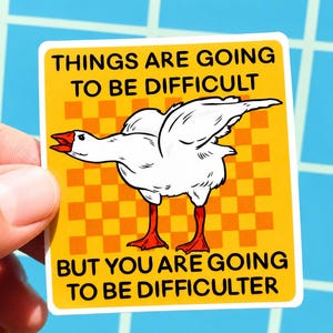 May include: A yellow sticker with the text "THINGS ARE GOING TO BE DIFFICULT BUT YOU ARE GOING TO BE DIFFICULTER." The sticker features a cartoon goose with white feathers, red feet, and a red beak. The background has an orange and white checkered pattern.