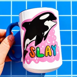 May include: White ceramic mug with a blue handle and rim. The mug features a black and white orca illustration on a pink background with the word "SLAY" in colorful bubble letters. The mug is held by a hand.