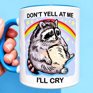 May include: A white ceramic mug with a blue handle and rim, featuring a graphic of a round, grey raccoon sitting under a colourful rainbow and yellow stars. The text above the raccoon reads DON'T YELL AT ME and below it reads I'LL CRY.