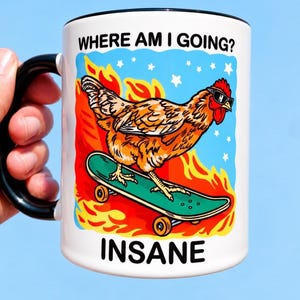 May include: A white ceramic mug with a black handle and rim featuring a graphic of a brown chicken wearing sunglasses on a green skateboard. The background shows orange flames and white stars against a blue sky. Text reads WHERE AM I GOING? and INSANE.