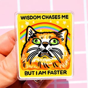 May include: A rectangular acrylic badge reel featuring an orange cat with wide green eyes and a surprised expression. A colorful rainbow and white stars are in the background. The text reads WISDOM CHASES ME BUT I AM FASTER in black.