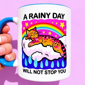 May include: A white ceramic mug with a bright blue handle and rim. The graphic features an orange tabby cat sleeping on a white cloud under a rainbow and yellow stars, with blue raindrops below. Text reads A RAINY DAY WILL NOT STOP YOU.