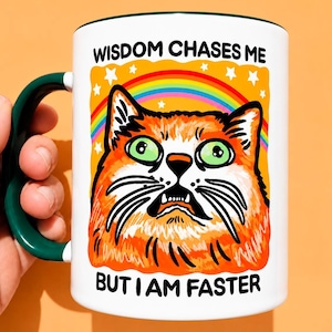 May include: A white ceramic mug with a dark green handle and rim. The front features an orange cat illustration with wide green eyes, a rainbow, and white stars. The text reads: WISDOM CHASES ME BUT I AM FASTER in black capital letters.