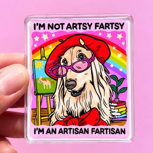 May include: A rectangular acrylic magnet featuring a golden dog wearing a red beret and pink glasses. The illustration includes a rainbow, an easel with a painting, and a stack of books. The text reads I'M NOT ARTSY FARTSY I'M AN ARTISAN FARTISAN.