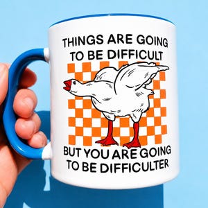 Puede incluir: Una taza de cerámica blanca con asa y borde azules. El frente presenta una ilustración de un ganso blanco con patas y pico rojos sobre un fondo de cuadros naranjas y blancos, con el texto: THINGS ARE GOING TO BE DIFFICULT BUT YOU ARE GOING TO BE DIFFICULTER.