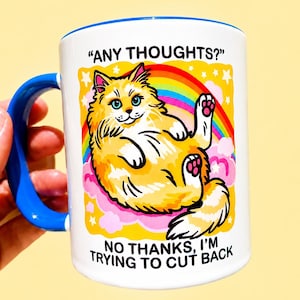 Puede incluir: Una taza de cerámica blanca con asa e interior azules. El frente presenta un gráfico de un gato naranja esponjoso acostado sobre nubes rosas bajo un arcoíris. El texto dice ANY THOUGHTS? arriba y NO THANKS, I'M TRYING TO CUT BACK en la parte inferior.
