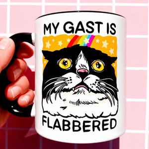 Op de afbeelding: Witte keramische mok met een zwarte handgreep en rand. De mok heeft een cartoonkat met grote gele ogen, zwart-witte vacht en de tekst "MY GAST IS FLABBERED" in het zwart. Een regenboog en sterren staan boven het hoofd van de kat.