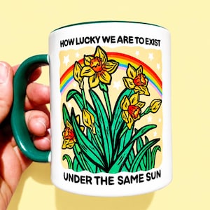 May include: A white ceramic mug with a dark green handle and rim. The front features a graphic of yellow daffodils, green leaves, and a rainbow against a pale yellow background with white stars. Text reads: HOW LUCKY WE ARE TO EXIST UNDER THE SAME SUN.