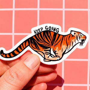 May include: A sticker featuring a running tiger with orange and black stripes. The sticker has the words "KEEP GOING" in black text above the tiger. The sticker is held by a hand against a pink grid background.