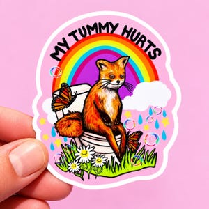 May include: A die-cut sticker featuring a fox sitting on a white toilet with the text MY TUMMY HURTS in black. The background includes a colorful rainbow, white clouds, blue raindrops, yellow stars, floating bubbles, and orange butterflies.
