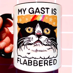May include: White ceramic mug with a black handle and rim. The mug features a cartoon cat with large yellow eyes, a rainbow, and the text "MY GAST IS FLABBERED" in black lettering. The cat is black and white.