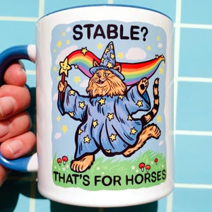 Op de afbeelding: Witte keramische mok met een blauw handvat en rand, met een cartoon kat tovenaar met een regenboog en de tekst "STABLE? THAT'S FOR HORSES." De kat draagt een blauwe jurk met gele sterren en een tovenaars hoed.