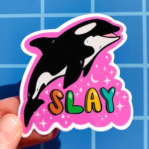 May include: A pink sticker featuring a black and white orca leaping from the water. The orca is surrounded by white stars and a crescent moon. The word "SLAY" is written in rainbow colours below the orca.