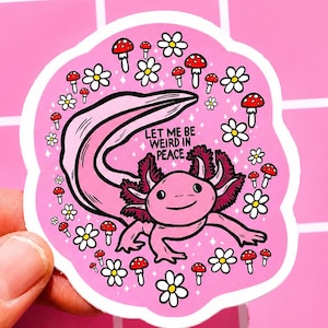 May include: A pink sticker featuring a cartoon axolotl with pink gills and a long tail, surrounded by white flowers and red-capped mushrooms. The sticker has the text "LET ME BE WEIRD IN PEACE" in black. The sticker is held by a hand.