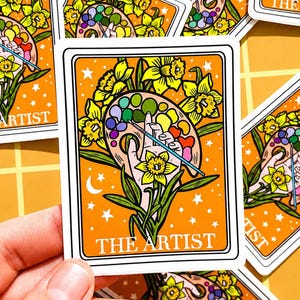 May include: Orange sticker featuring a hand holding a paint palette with a paintbrush, surrounded by yellow daffodils. The text "THE ARTIST" is at the bottom. Stars and a crescent moon decorate the background. The sticker has a white border.