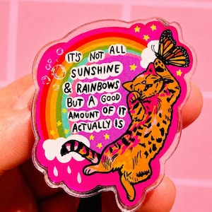 May include: An acrylic badge reel featuring an orange tabby cat reaching for a butterfly. The design includes a rainbow, white clouds, and the text: IT'S NOT ALL SUNSHINE & RAINBOWS BUT A GOOD AMOUNT OF IT ACTUALLY IS, set against a bright pink background.