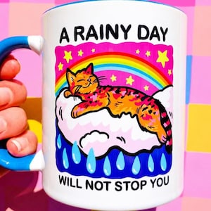 May include: White ceramic mug with a blue handle and rim. The mug features a colorful illustration of a cat resting on a cloud under a rainbow, with the text "A RAINY DAY WILL NOT STOP YOU".